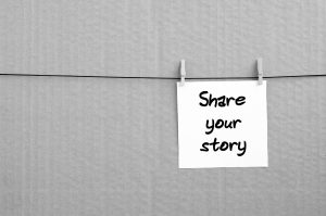 Share your story. Note is written on a white sticker that hangs with a clothespin on a rope on a background of brown cardboard